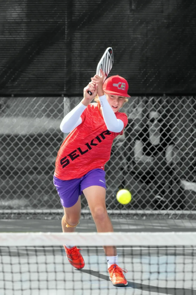 young boy wearing red pickleball shirt and cap in mid-swing playing pickleball