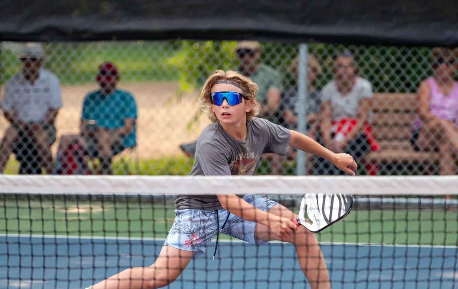 young boy with blonde hair wearing colored reflective sunglasses playing pickleball